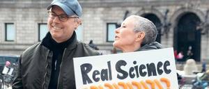 Eine Frau steht auf der Straße mit einem Schild auf der Hand, darauf steht: Real Science, Fake President.