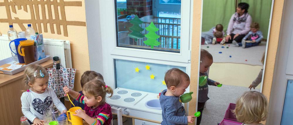 Kleinkinder spielen in einer Kita mit einem Spielzeug-Kochherd. Im Hintergrund sitzt eine Erzieherin mit anderen Kindern zusammen.
