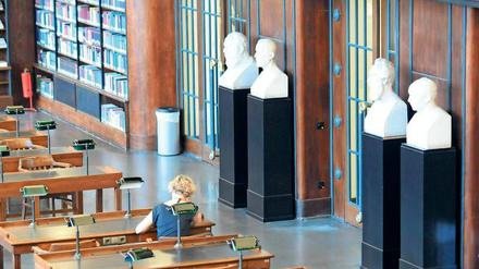 Eine Frau sitzt in einer Bibliothek an einem Tisch, im Hintergrund sind Gipsköpfe von Gelehrten zu sehen.