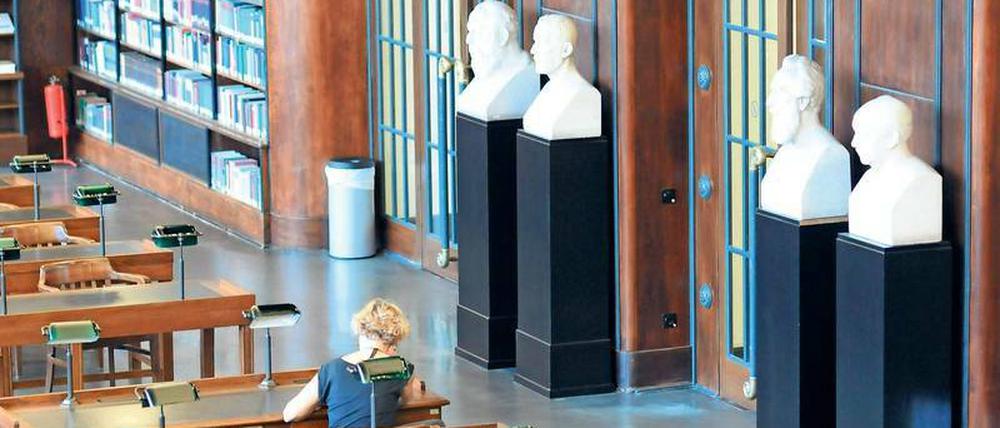 Eine Frau sitzt in einer Bibliothek an einem Tisch, im Hintergrund sind Gipsköpfe von Gelehrten zu sehen.