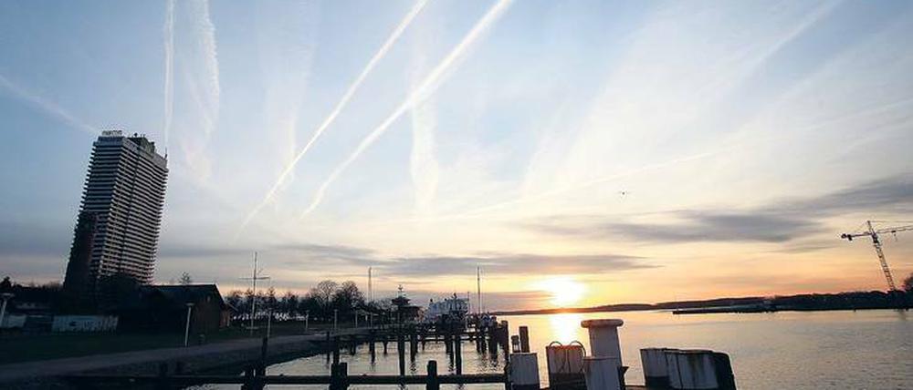 Das Bild zeigt einen Sonnenuntergang hinter einer Hafenanlage, über einem scheinbar schräg stehenden Hochhaus sind strahlenförmige Kondensstreifen am Himmel zu sehen.