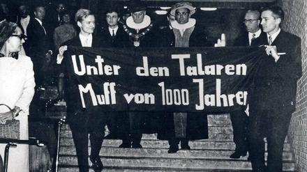 Zwei junge Männer tragen ein Plakat mit der Aufschrift "Unter den Talaren Muff von 1000 Jahren", dahinter gehen zwei Männer in akademischen Roben.