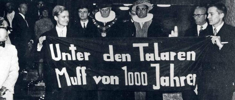 Zwei junge Männer tragen ein Plakat mit der Aufschrift "Unter den Talaren Muff von 1000 Jahren", dahinter gehen zwei Männer in akademischen Roben.