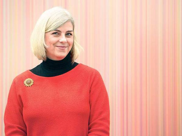 Eine Frau in einem roten Pullover steht lächelnd vor einer gestreiften Tapete.