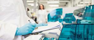 Eine Labormitarbeiterin im weißen Kittel und mit blauen Handschuhen schreibt mit einem Stift auf einem Klemmbrett.