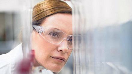 Eine junge Frau mit Schutzbrille ist hinter gläsernen Laborgeräten zu sehen.