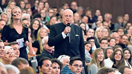 Ein Mann mit einem Mikrofon in der Hand steht inmitten von Studierenden, die in einem Hörsaal sitzen.