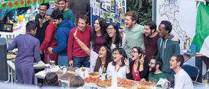 Studierende stehen hinter einem Buffet mit Essensangeboten und lachen winkend in die Kamera.