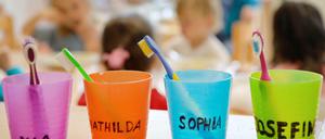 Zahnbürsten und Zahnputzbecher mit den Vornamen der Kinder stehen in einer Kindertagesstätte auf einem Tisch. 