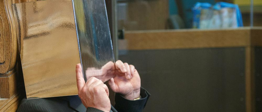 Der Angeklagte sitzt auf der Anklagebank und hält sich einen Aktenordner vor das Gesicht. 