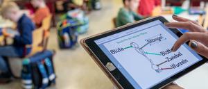 Eine Lehrkraft steht im Klassenraum und tippt auf ein Schaubild auf einem Tablet.