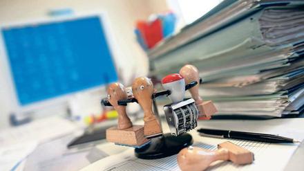 Blick auf einen Behördenschreibtisch - mit Stempeln, Aktenberg und Computerbildschirm.