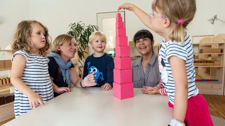 Kitakinder bauen einen Turm aus Klötzchen, zwei Erzieherinnen schauen zu.