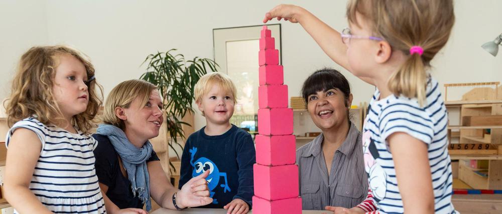 Kitakinder bauen einen Turm aus Klötzchen, zwei Erzieherinnen schauen zu.