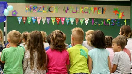 Erstklässler in einer Grundschule sitzen auf einer Bank mit dem Rücken zur Kamera. Da drüber ein "Herzlich Willkommen"-Transparent.