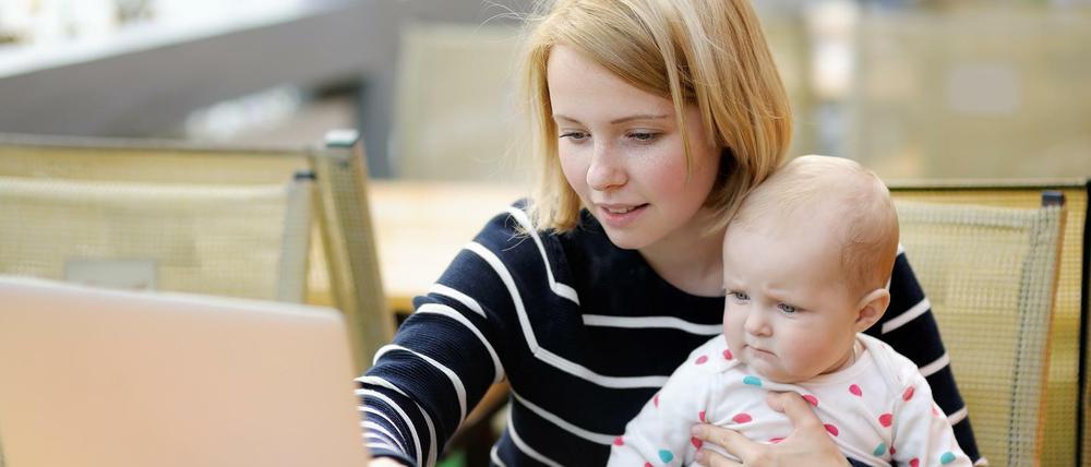 Eine junge Mutter sitzt mit ihrem Baby auf dem Schoß vor einem Laptop. (Symbolbild)