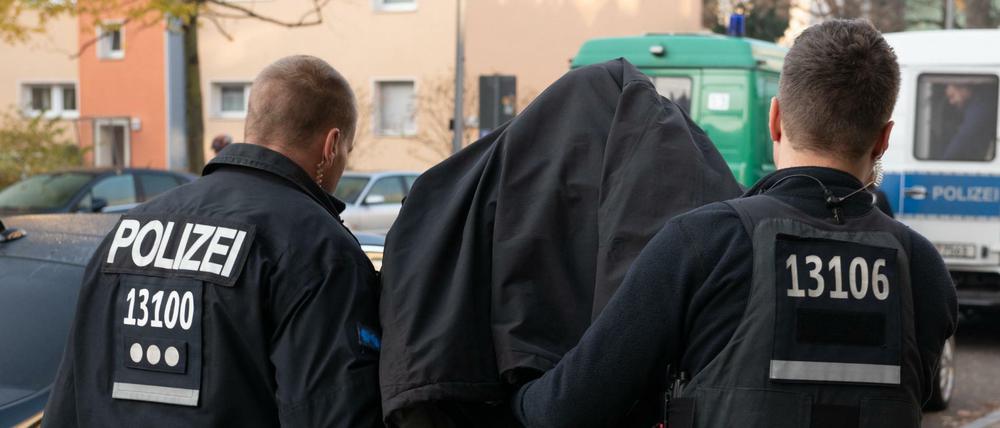 Polizeibeamte führen mit Handschellen einen festgenommenen Mann nach einer Razzia in einem Wohnhaus in Tempelhof ab.