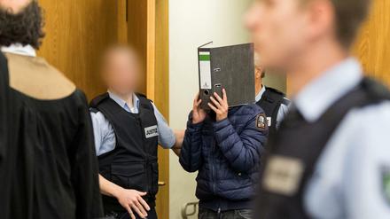 Einer der Angeklagten kommt mit einem Aktenordner vor dem Gesicht in den Gerichtssaal. 