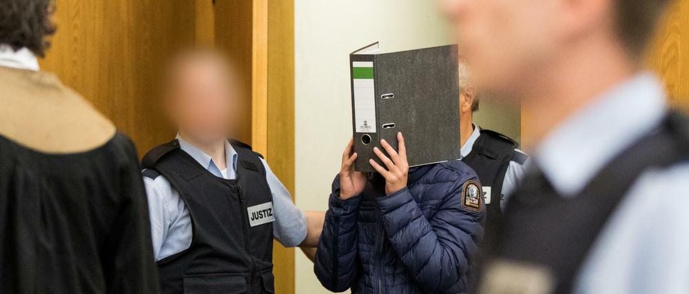 Einer der Angeklagten kommt mit einem Aktenordner vor dem Gesicht in den Gerichtssaal.