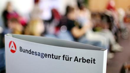 Ein Schild mit der Aufschrift "Bundesagentur für Arbeit" steht einem vollen Wartezimmer im Jobcenter. 