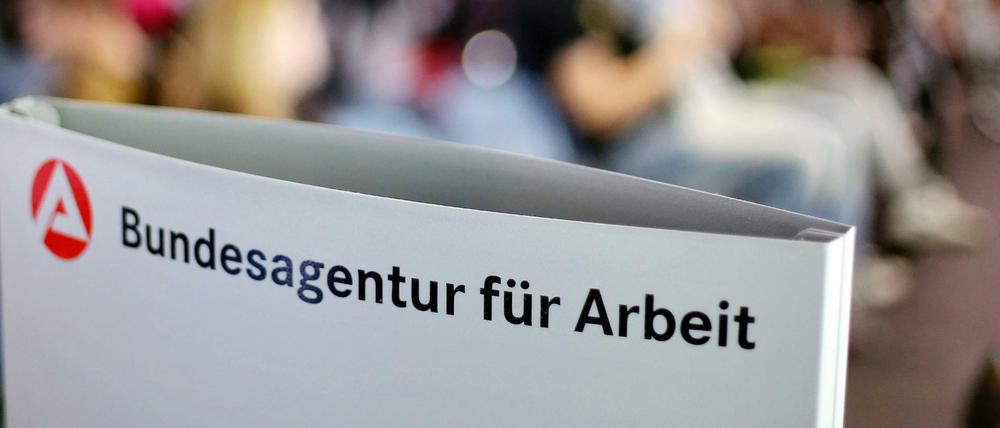 Ein Schild mit der Aufschrift "Bundesagentur für Arbeit" steht einem vollen Wartezimmer im Jobcenter.