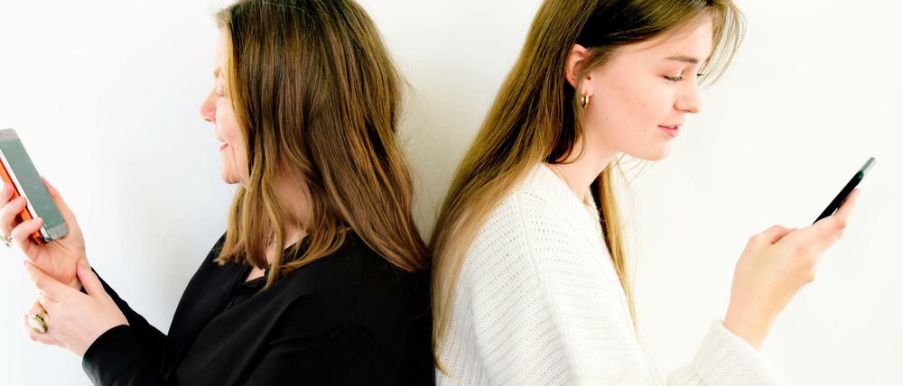 Zwei Frauen sitzen Rücken an Rücken und tippen Sprachnachrichten in ihre Handys ein.