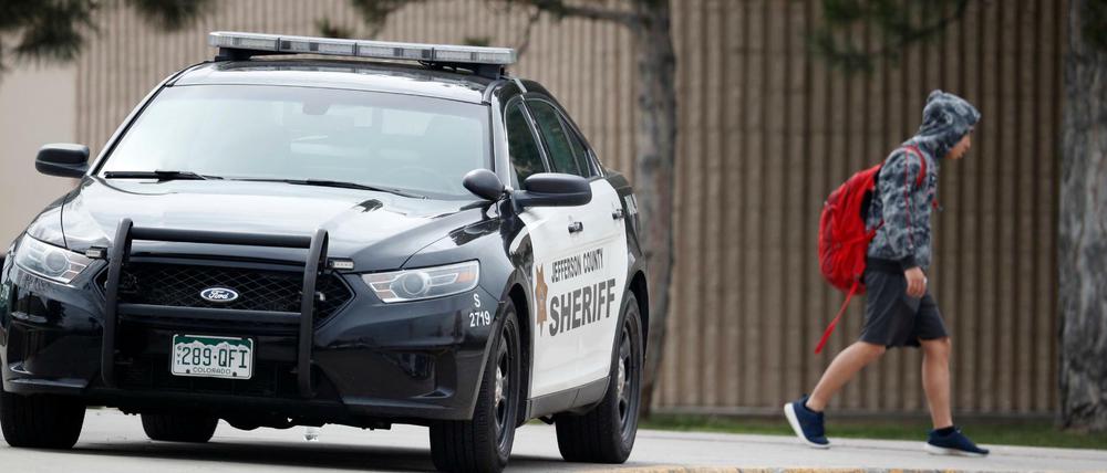 Ein Polizeiauto steht vor der Columbine High School, während Schüler diese verlassen.