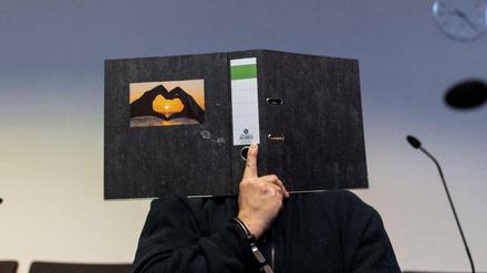 Der 58-jährige Angeklagte sitzt im Gerichtssaal und hält einen Aktenordner mit einem Herz aus zwei Händen vor sein Gesicht.