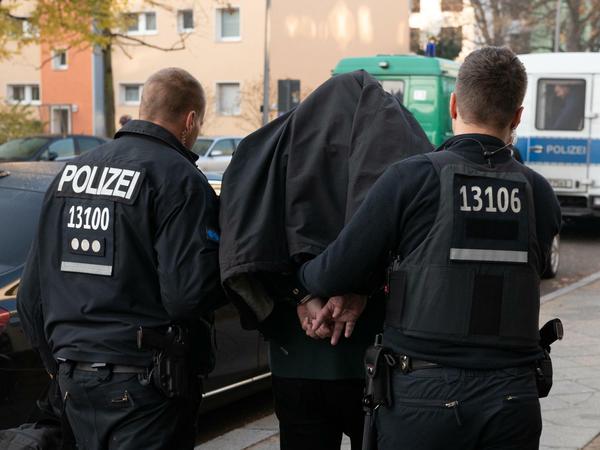 Polizeibeamte führen mit Handschellen einen festgenommenen Mann nach einer Razzia in einem Wohnhaus in Tempelhof ab.
