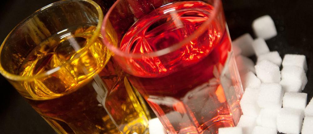 Drei Gläser gefüllt mit roter Limonade, Cola und einem Energy-Drink stehen neben Zuckerwürfeln auf einem Tisch. 