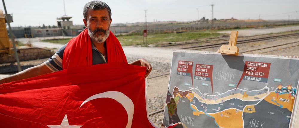 Ein Mann mit einer türkischen Flagge steht an der Grenze zwischen der Türkei und Syrien neben einer Karte, welche die geplante militärische Operation der Türkei in Syrien zeigt.