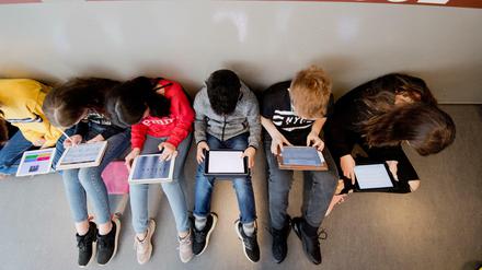 Schüler und Schülerinnen sitzen an einer Wand auf dem Fußboden und arbeiten mit Tablet-Computern.