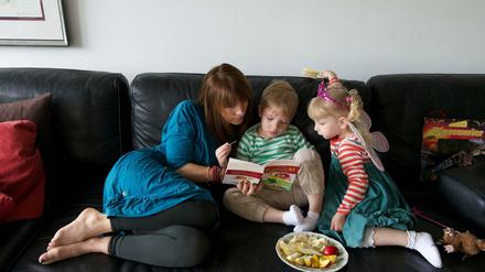 Eine Frau sitzt mit zwei Kindern auf einem Sofa und schaut mit ihnen in ein Buch; ein Obstteller steht bereit.