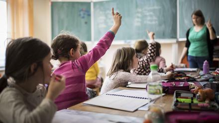 Grundschulkinder sitzen in einem Klassenraum und melden sich, die Lehrerin hebt auch den Finger.