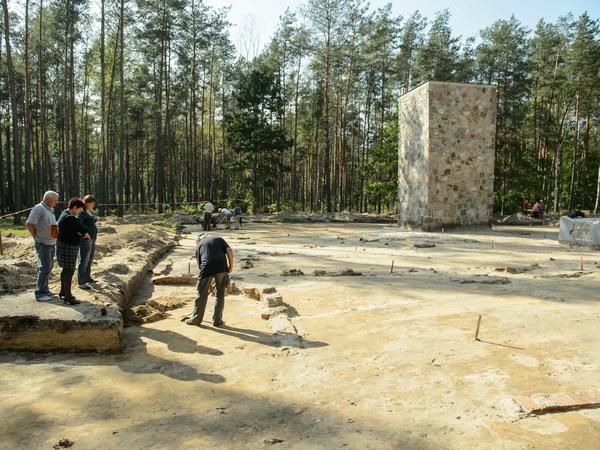 Archäologische Arbeiten auf einem Freigelände am Waldrand, im Hintergrund ist ein gemauertes Mahnmal zu sehen.
