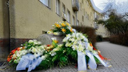 An einer Hausecke ist ein Gedenkstein mit Trauergestecken bedeckt.