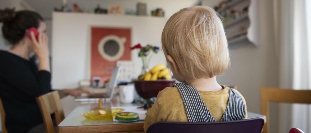 Eine Frau sitzt im Homeoffice an ihrem Laptop und telefoniert, während ihr Kind neben ihr in einem Kinderstuhl am Tisch sitzt.