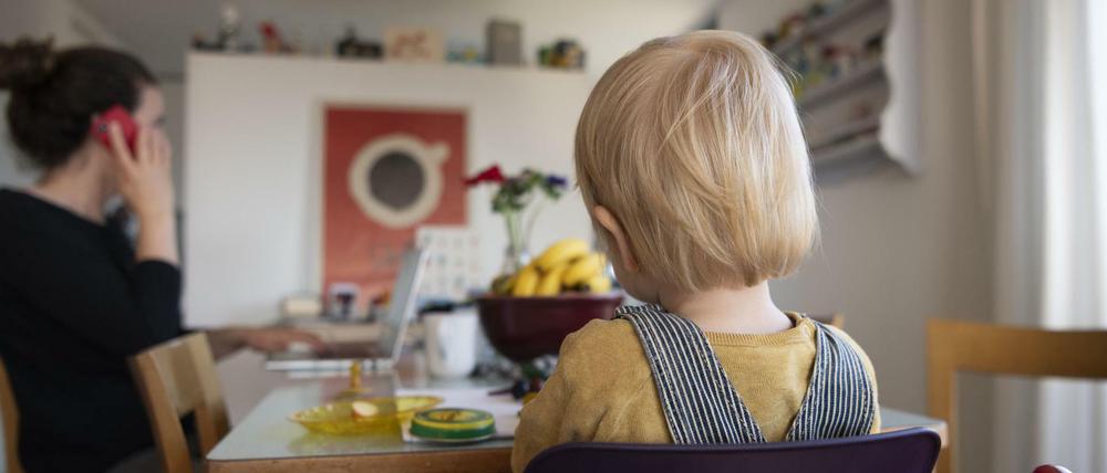 Eine Frau sitzt im Homeoffice an ihrem Laptop und telefoniert, während ihr Kind neben ihr in einem Kinderstuhl am Tisch sitzt.