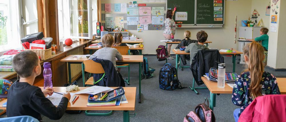 Grundschüler sitzen in einem Klassenraum an Einzeltischen, ihre Lehrerin erklärt etwas an der Schiefertafel.