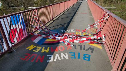 Auf einer Brücke liegt rot-weißes Absperrband auf dem Schriftzug Willkommen, Bienvenue.