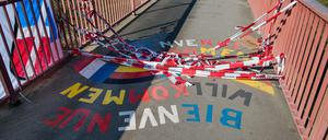 Auf einer Brücke liegt rot-weißes Absperrband auf dem Schriftzug Willkommen, Bienvenue.