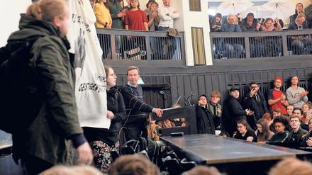 Bernd Lucke steht in einem Hörsaal am Rednerpult, umgeben von protestierenden Studierenden und Aktivisten.