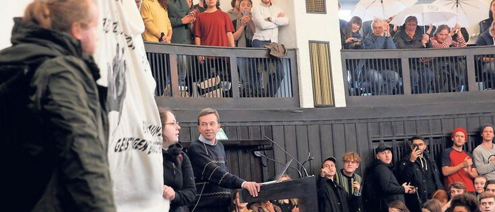 Bernd Lucke steht in einem Hörsaal am Rednerpult, umgeben von protestierenden Studierenden und Aktivisten.