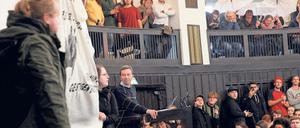 Bernd Lucke steht in einem Hörsaal am Rednerpult, umgeben von protestierenden Studierenden und Aktivisten.