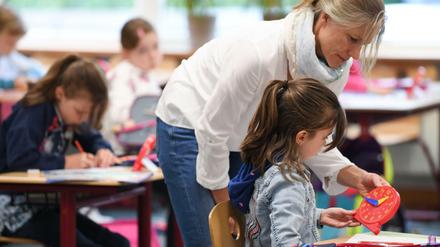 Eine Grundschullehrerin beugt sich über eine Zweitklässlerin und erklärt ihr die Uhrzeit an einer Holzuhr.