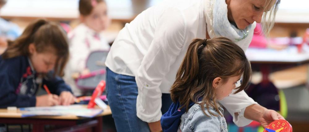Eine Grundschullehrerin beugt sich über eine Zweitklässlerin und erklärt ihr die Uhrzeit an einer Holzuhr.