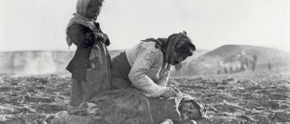Auf einem unbestellten Feld beugt sich eine Frau über ein am Boden liegendes Kind, ein zweites Kind steht andächtig daneben.