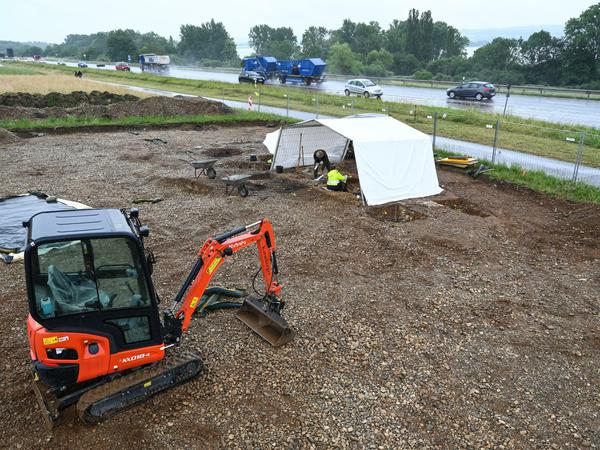 Neben einer archäologischen Fundstätte - von einem Zelt überspannt - fahren Autos auf einer Bundesstraße, im Vordergrund steht ein kleiner Bagger.