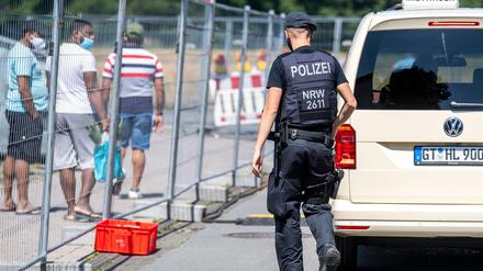 Ein Polizist geht zu einer Gruppe von Menschen, die hinter einem Bauzaun unter Quarantäne stehen. Nach dem Corona-Ausbruch beim Fleischverarbeiter Tönnies verhängen die Behörden einen Lockdown auch für den Kreis Gütersloh und den Nachbarkreis.