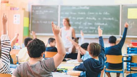 In einem Klassenraum sitzen Schüler an ihren Tischen und melden sich, vorne steht eine Lehrerin.